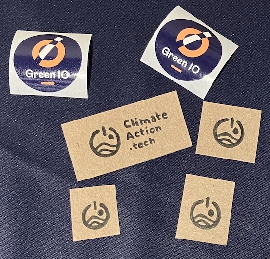 Close-up of square cardboard badges, with the Climate Action Tech logo hand-drawn. And Green IO stickers with their logo